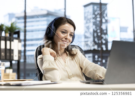 Joyful female CEO working on her business and listening to podcast, using headset and multitasking in smart urban office. Ambitious entrepreneur having fun with an interesting audio documentary. 125356981