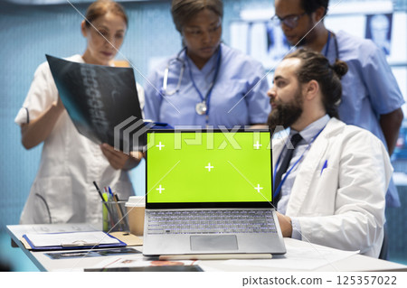 Team of doctors consulting analytics next to green screen on laptop in medical cabinet, examining x ray scan results to find the diagnosis. Licensed medics review hospital records at desk. 125357022