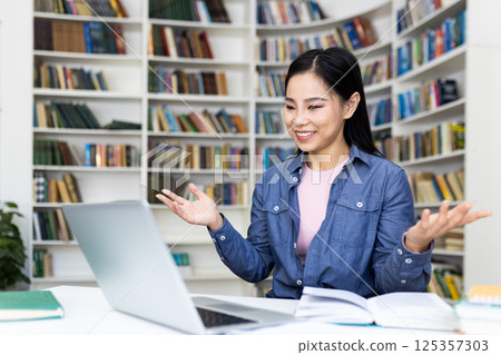 A smiling asian woman sits at a desk in a library, gesturing while looking at her laptop. Bookshelves filled with books surround her. A smiling asian woman sits at a desk in a library, gesturing while looking at her laptop. Bookshelves filled with books surround her. 125357303