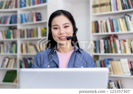 An attractive asian woman smiles while wearing a headset, working or studying remotely. The background shows bookshelves. 125357307