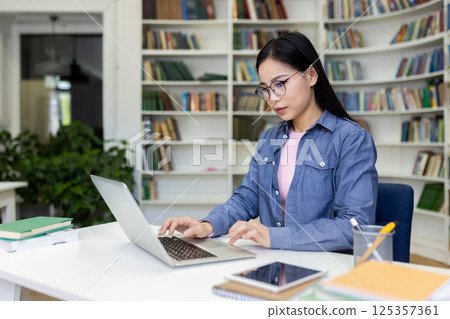 An asian woman, wearing glasses, works on a laptop at a library table, surrounded by books. She is focused on her task in a study setting. 125357361