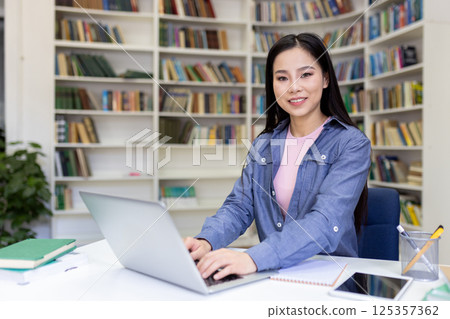 A smiling Asian woman works on her laptop at a desk in a library, surrounded by books and a focused environment. 125357362