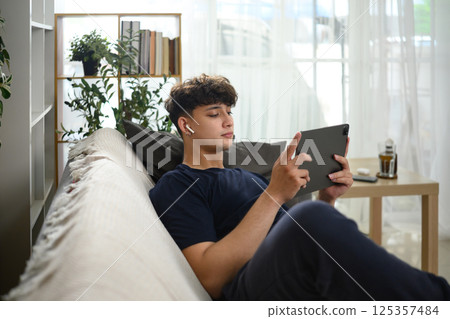 Young man lounging on couch with earbuds and watching content on a digital tablet Young man lounging on couch with earbuds and watching content on a digital tablet 125357484