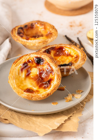 Three Pastel de Nata, Portuguese custard tarts, are presented on a plate with cinnamon and vanilla beans. 125358079