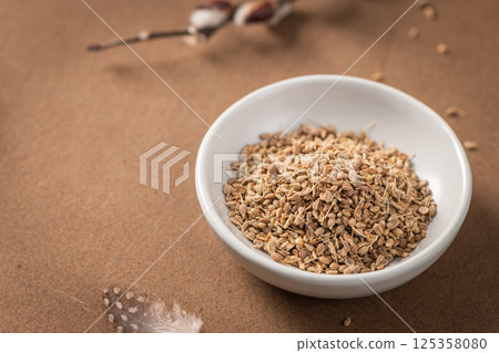 Fennel seeds in a white bowl on a textured brown surface with a delicate feather and blurred plant stem in the background 125358080