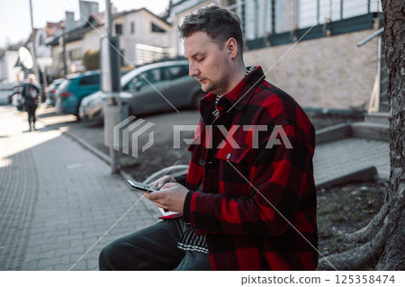 Male student with smartphone wearing plaid shirt sitting on bench while waiting for transport at tram or bus station. Male student with smartphone wearing plaid shirt sitting on bench while waiting for transport at tram or bus station. 125358474
