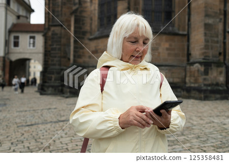 Happy senior 50s 60s woman holding backpack on shoulder sightseeing in old town of Praha, looking at map in a smartphone enjoying travel tourism and freedom. Happy senior 50s 60s woman holding backpack on shoulder sightseeing in old town of Praha, looking at map in a smartphone enjoying travel tourism and freedom. 125358481