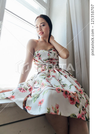 Beautiful brunette woman in summer dress with flowers sitting by the window Beautiful brunette woman in summer dress with flowers sitting by the window 125358707