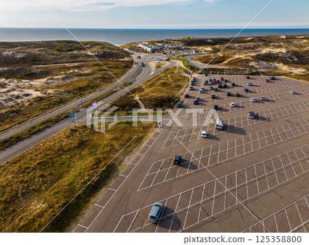 Aerial view of a coastal parking lot near sandy dunes with scattered cars, clear blue sky, and the sea in the distance. A road winds through the landscape leading to a beachside area. Aerial view of a coastal parking lot near sandy dunes with scattered cars, clear blue sky, and the sea in the distance. A road winds through the landscape leading to a beachside area. 125358800