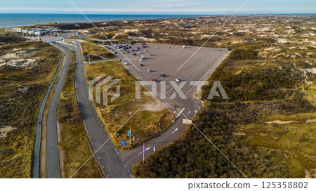 Aerial view of a coastal parking lot near sandy dunes with scattered cars, clear blue sky, and the sea in the distance. A road winds through the landscape leading to a beachside area. Aerial view of a coastal parking lot near sandy dunes with scattered cars, clear blue sky, and the sea in the distance. A road winds through the landscape leading to a beachside area. 125358802