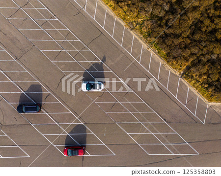 Aerial view of a large, organized parking lot filled with colorful cars. Bright sunlight casts long shadows, highlighting the neat rows and road markings surrounding the area. 125358803