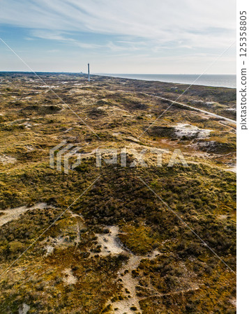 Aerial view of a coastal dune landscape with a lighthouse in the distance, sandy paths winding through the terrain, and the sea stretching out under a calm, partly cloudy sky. 125358805