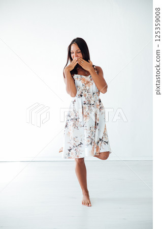 Brunette in summer dress covers her mouth with her hand in white room 125359008