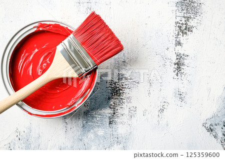 Red paintbrush resting on open can of red paint over textured white and gray painted surface. 125359600