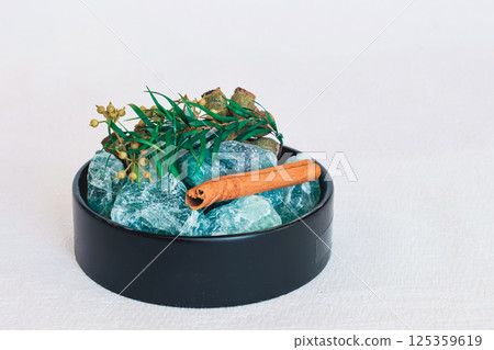 The photo shows black dishe containing various natural ingredients. In the center of the bowl is a stick of cinnamon, and around it are pieces of green crystal 125359619
