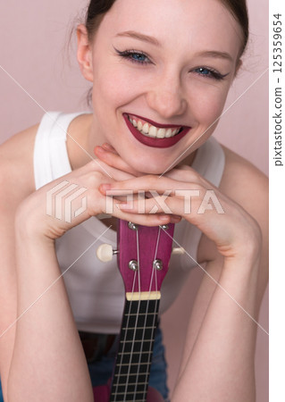 Portrait of young woman musician with ukulele. Beautiful 18 year old woman with big, toothy smile, positive attitude and joyful expression. She's head resting on her hands, cradling ukulele vertically Portrait of young woman musician with ukulele. Beautiful 18 year old woman with big, toothy smile, positive attitude and joyful expression. She's head resting on her hands, cradling ukulele vertically 125359654