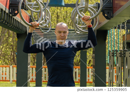 Muscular man doing pull-ups and gymnastic ring exercises on sports ground. Summer. Outdoor sports public park. Healthy lifestyle. Close up. 125359905