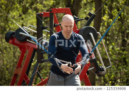 Man performing exercise in crossover on outdoor fitness equipment, focusing on his arm strength. Bodybuilding. Outdoor sport in public park. Summer season. 125359906