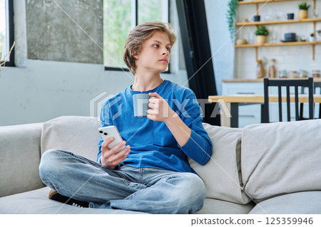 Relaxed teenager guy sitting at home on the couch with smartphone and cup of tea 125359946