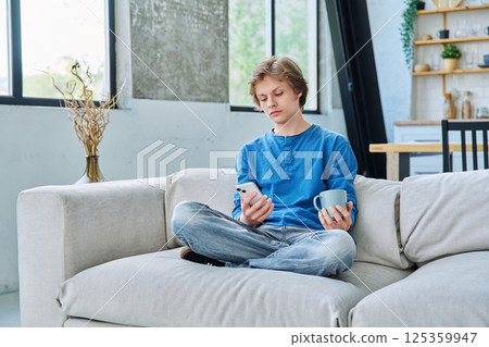 Relaxed teenager guy sitting at home on the couch with smartphone and cup of tea 125359947
