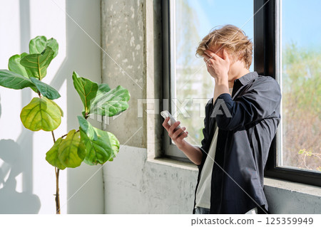 Upset sad teenager guy looking at smartphone screen, at home near window Upset sad teenager guy looking at smartphone screen, at home near window 125359949