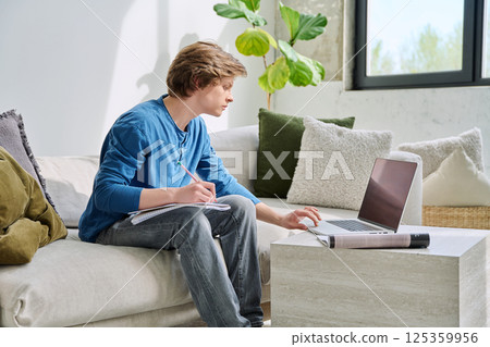 Teenager guy student studying sitting at home on sofa using laptop textbook notebook Teenager guy student studying sitting at home on sofa using laptop textbook notebook 125359956