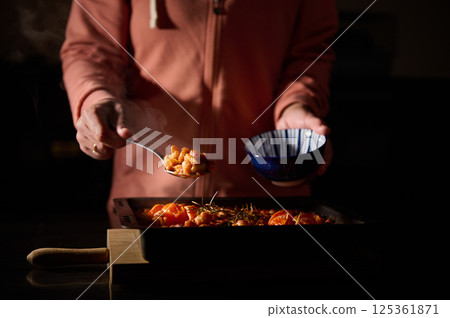 A person serves freshly cooked baked beans garnished with herbs into a blue bowl, captured in a dimly-lit kitchen setting emphasizing deliciousness and care in meal preparation. 125361871