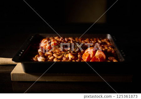 A tray of savory oven-baked beans garnished with tomatoes and rosemary, presented in warm, ambient lighting. The rich texture and flavors evoke an inviting homemade, rustic feel. 125361873