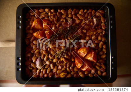 Close-up of oven-baked beans garnished with tomatoes, garlic, and fresh rosemary, highlighting a rustic, homemade meal perfect for cozy dining. 125361874