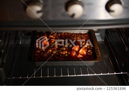 A baking tin filled with baked beans and tomatoes, flavored with rosemary, inside a preheated oven. The warm lighting enhances the inviting and appetizing nature of the dish during preparation. 125361896