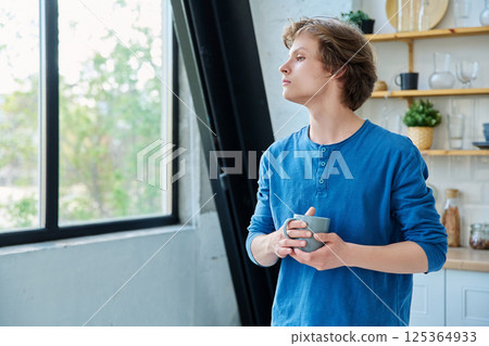 Teenage serious guy with cup in hands in home interior, looking out window Teenage serious guy with cup in hands in home interior, looking out window 125364933
