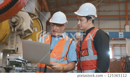 Engineer teaching apprentice to use metal sheet stamping machine, industrial team is working on various projects with laptop computer, worker work together to check the operation system in a factory 125365112