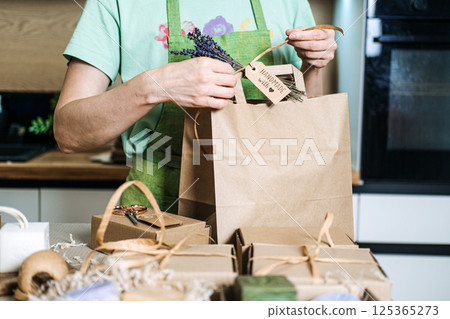 A small business owner ties a Handmade with love tag and lavender sprig to a brown paper bag surrounded by gift boxes. Home-based business, handmade goods 125365273