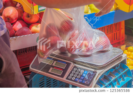 Transparent plastic bags with ripe fruits of red peaches, apples, which seller at market lays out on surface of measuring part of electronic floor scale. Trade, purchase of farm products for nutrition Transparent plastic bags with ripe fruits of red peaches, apples, which seller at market lays out on surface of measuring part of electronic floor scale. Trade, purchase of farm products for nutrition 125365586