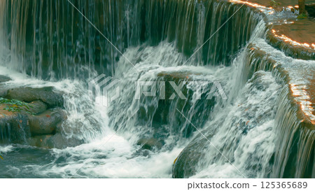 Close-up view of cascading waterfall flowing over layered rock formations in lush forest; movement of water and the beauty of natural landscapes 125365689