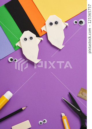 Origami paper ghosts with googly eyes with craft supplies on a purple background. Paper sheets, scissors, markers, glue and knife. Copyspace 125365757
