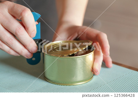 Womans hands open tin can with an can opener. Canned food and special knife 125366428