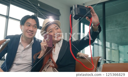 Wait boarding flight. Asian beautiful young loving couple smiling making selfie by their mirrorless camera and holding passports tickets while waiting for boarding in terminal hall airport Wait boarding flight. Asian beautiful young loving couple smiling making selfie by their mirrorless camera and holding passports tickets while waiting for boarding in terminal hall airport 125366829
