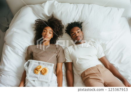 A man and a woman are laying on a bed with a newspaper 125367913