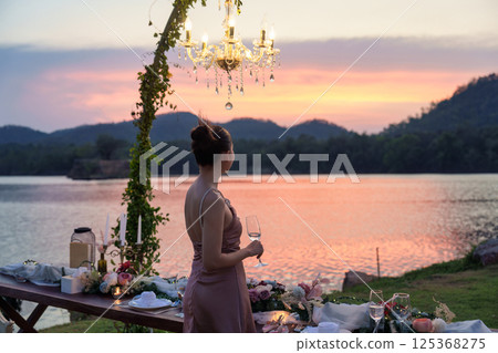 Beautiful asian woman standing holding a wine glass next to romantic dining table in the sunset 125368275