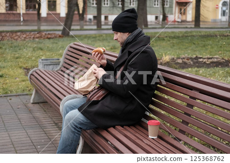 Pensive hipster drinking coffee sitting on a bench in the urban park. Handsome man wearing black coat eating street food, hot dog with paper cup in hand in the city. Stylish bearded middle-aged male. Pensive hipster drinking coffee sitting on a bench in the urban park. Handsome man wearing black coat eating street food, hot dog with paper cup in hand in the city. Stylish bearded middle-aged male. 125368762