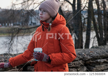 Teenager Girl Walking Alone on City Street. Portrait of 14 years old girl wearing eyeglasses, wool hat and winter jacket walks around the city. Urban Style. Rainy mood weather. Cold season. One person 125368804