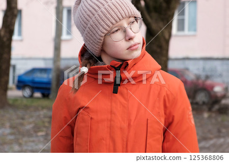 Teenager Girl Walking Alone on City Street. Portrait of 14 years old girl wearing eyeglasses, wool hat and winter jacket walks around the city. Urban Style. Rainy mood weather. Cold season. One person 125368806