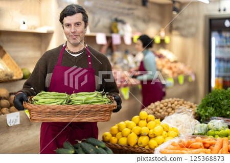 Man salesperson carries large basket of beans in pods from warehouse to sales area in shop Man salesperson carries large basket of beans in pods from warehouse to sales area in shop 125368873