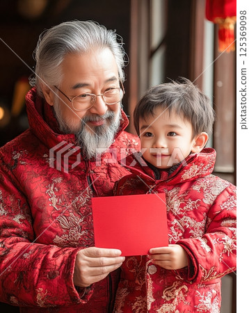 A heartwarming moment of a grandfather and grandson in matching red Chinese attire holding a red envelope, symbolizing blessings, prosperity, and family unity during the Chinese New Year celebration. 125369098