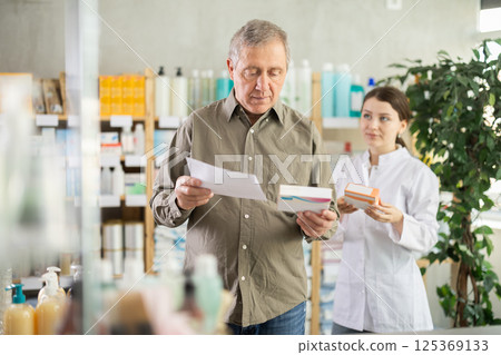 Senior man with doctors prescription selects pills,pharmacist in background Senior man with doctors prescription selects pills,pharmacist in background 125369133