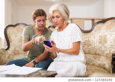 Wife and husband couple using smart phone watching videos, social media, reading news on cellphone at home 125369240