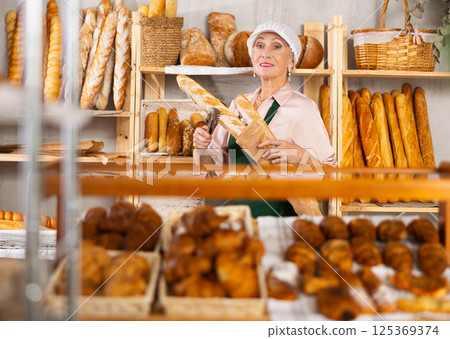 Elderly female bakery salesperson offers to buy bread, baguettes and croissants Elderly female bakery salesperson offers to buy bread, baguettes and croissants 125369374