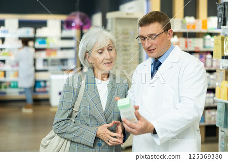 Man apothecary hold package with cream and talks with senior woman client about nuances of care Man apothecary hold package with cream and talks with senior woman client about nuances of care 125369380