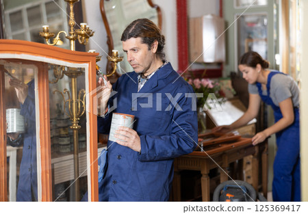 middle-aged furniture maker applies varnish with brush to wooden parts of vintage cabinet 125369417